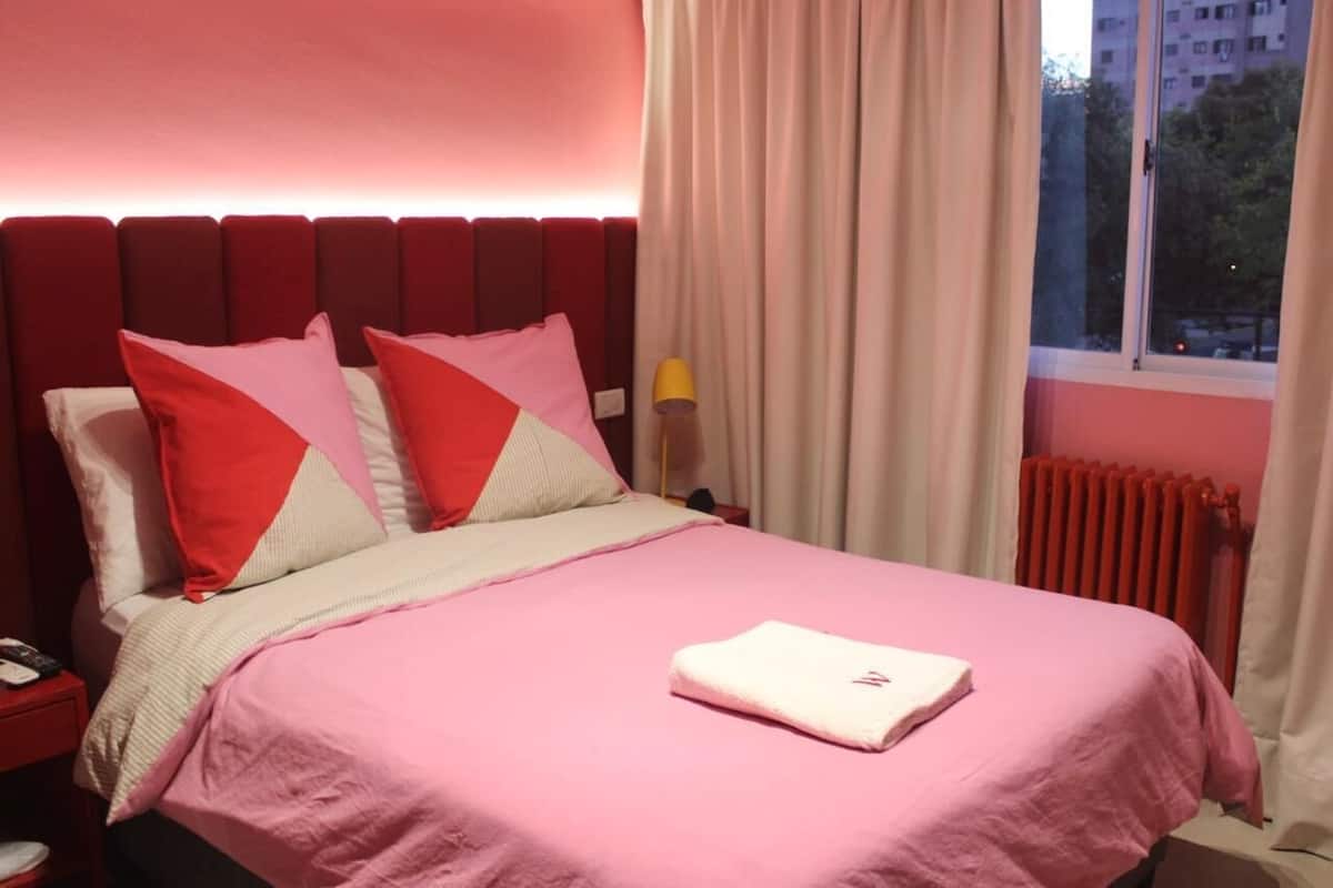 A bedroom with a pink bedspread, red and white pillows, a white blanket, and a nightstand with a phone and a lamp.
