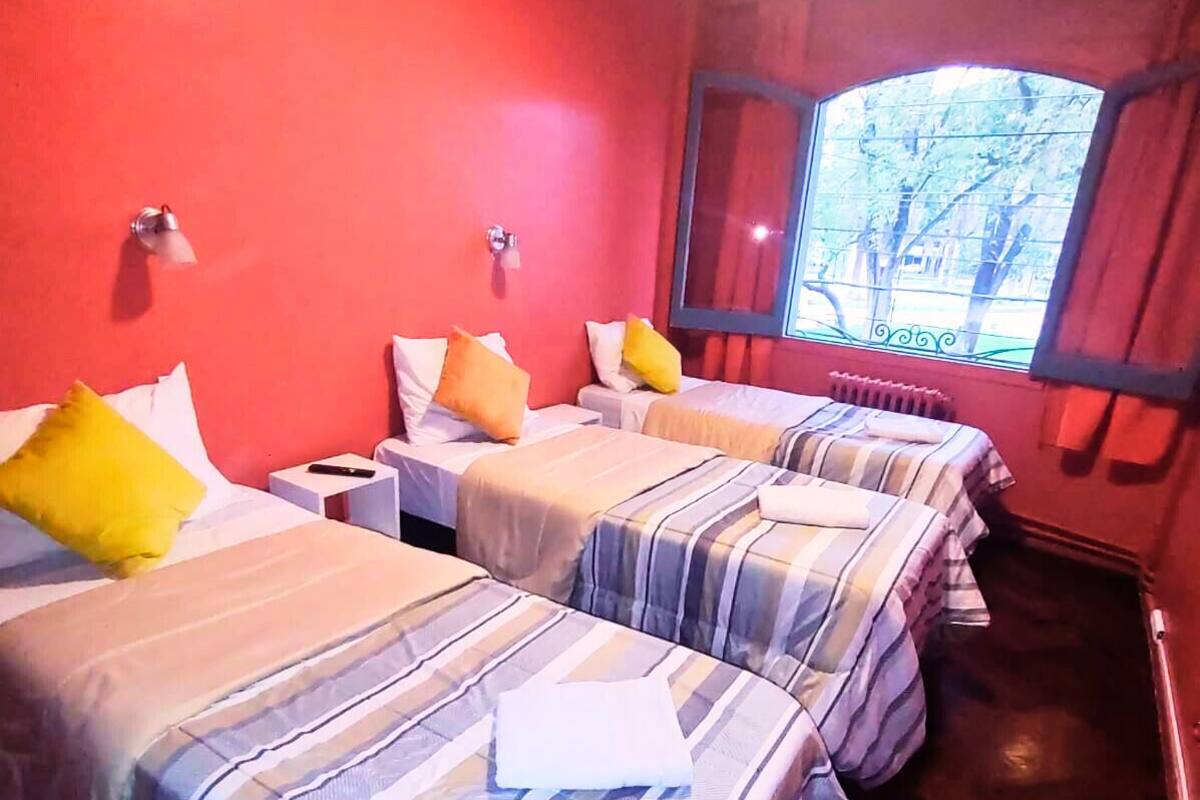 A hotel room with three beds, each with striped bedding and pillows, a window with a view of trees, and a red wall.