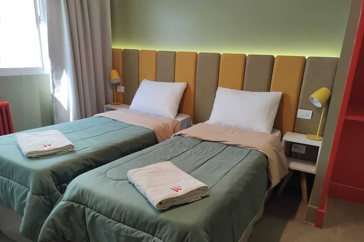A hotel room with two beds, each with a green bedspread and white pillows, and a headboard with vertical wooden panels.