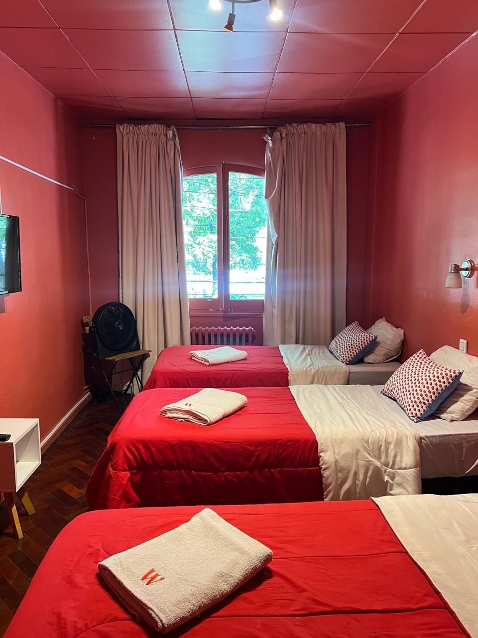 A hotel room with two beds, a window with curtains, a radiator, a fan, and a wall-mounted light fixture.