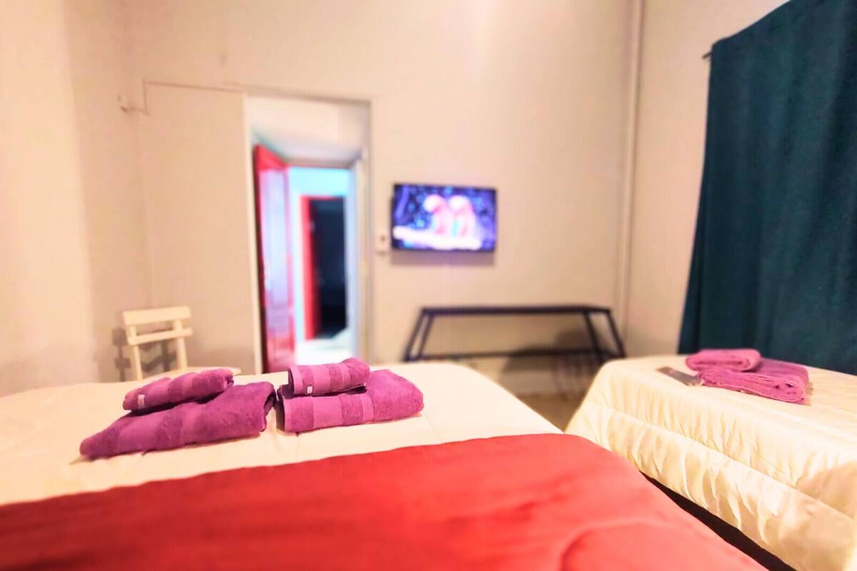 A hotel room with two beds, one with a red blanket and the other with a white one. There is a television on the wall and a door leading to another room.