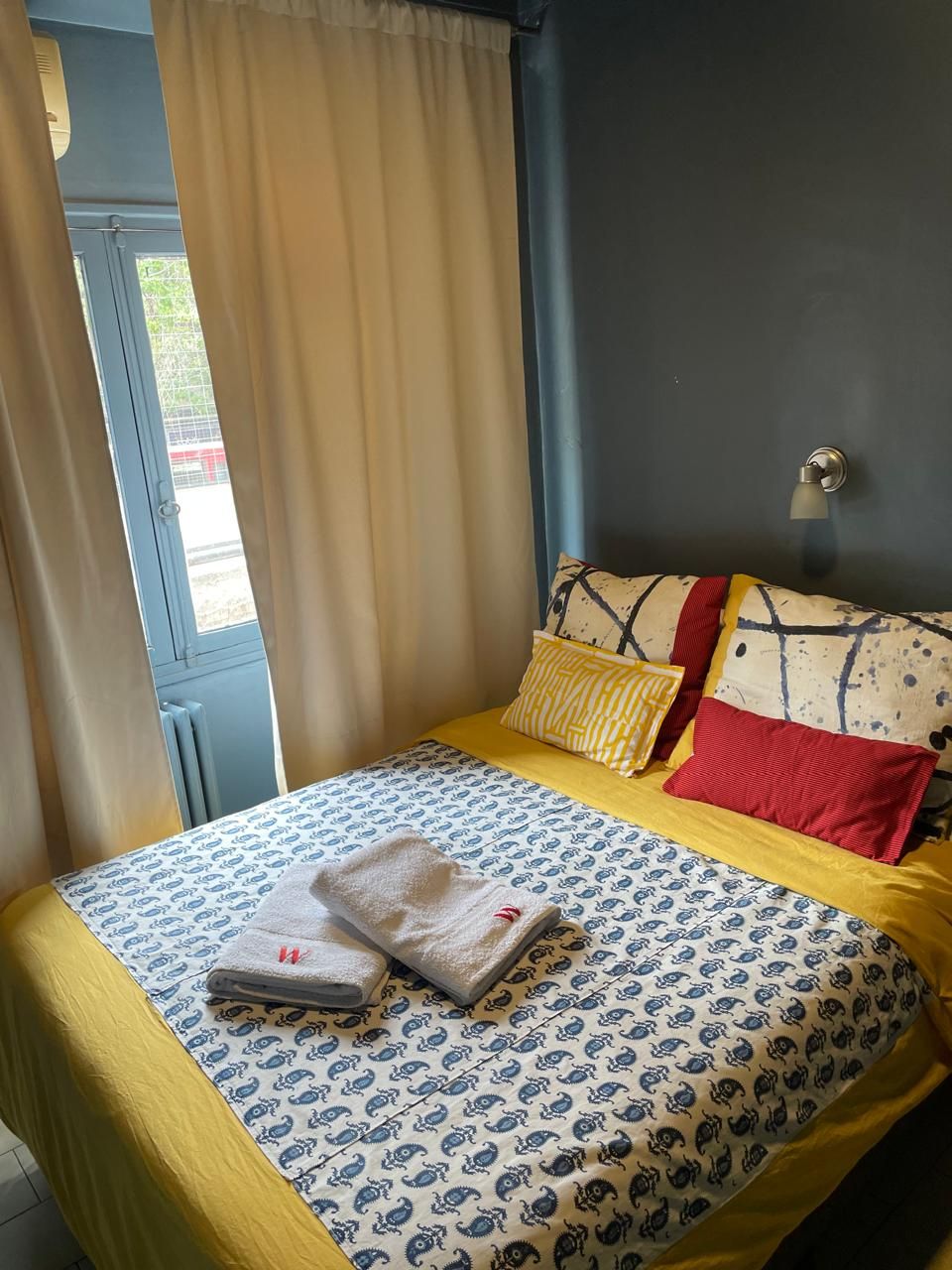 A bed with patterned bedding, a window with curtains, and a wall-mounted light fixture.