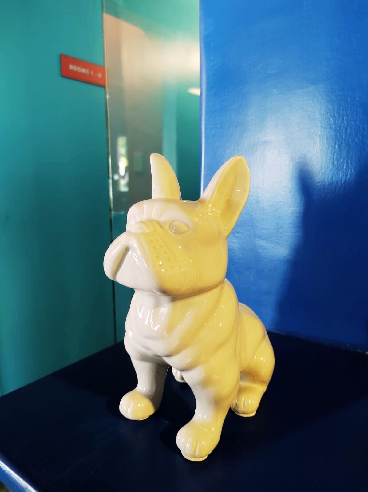 En keramisk hundefigur på en mørkeblå overflade, med en turkis væg og et rødt skilt i baggrunden.