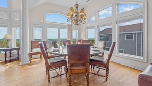 Une salle à manger spacieuse, avec une table ronde et six chaises, de grandes fenêtres offrant une vue sur l’océan et un lustre.