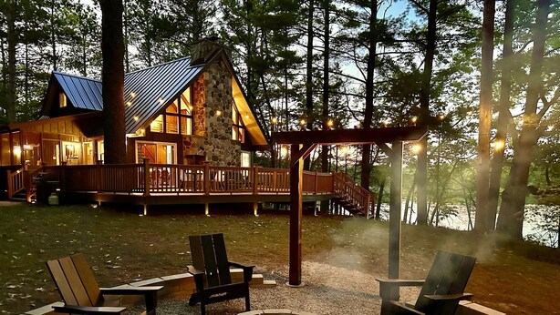 Una cabaña con chimenea de piedra, mobiliario exterior y una terraza con vistas a un cuerpo de agua.
