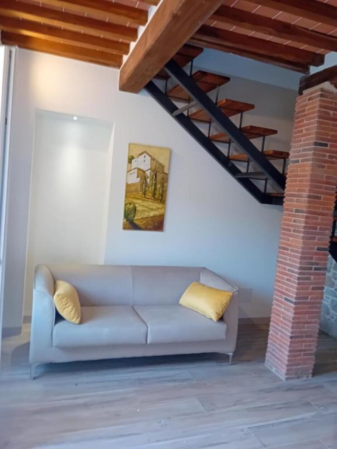 A modern living room with a light gray sofa, a painting of a rural scene, a brick fireplace, and a staircase with a metal railing.