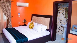 A hotel room with an orange wall, a bed with white bedding, a nightstand with a lamp, and a bathroom visible through an open door. - Annes luxury suites  (Lagos)