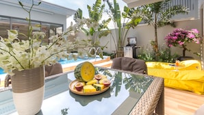 Uma mesa com tampo de vidro, um prato de frutas, um vaso de planta e vista para piscina e área de estar externa.