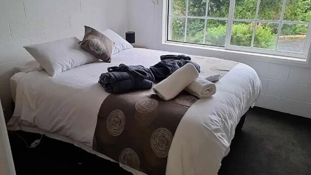 A neatly made bed with a brown patterned bedspread and white pillows, a rolled towel, and a black robe on the bed. - Orchard Apartment Devonport (Don)