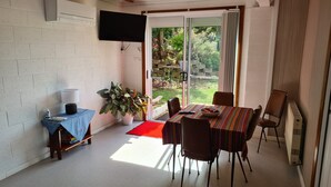 A dining area with a table and chairs, a television mounted on the wall, and a view of the garden through a glass door. - Orchard Apartment Devonport (Don)