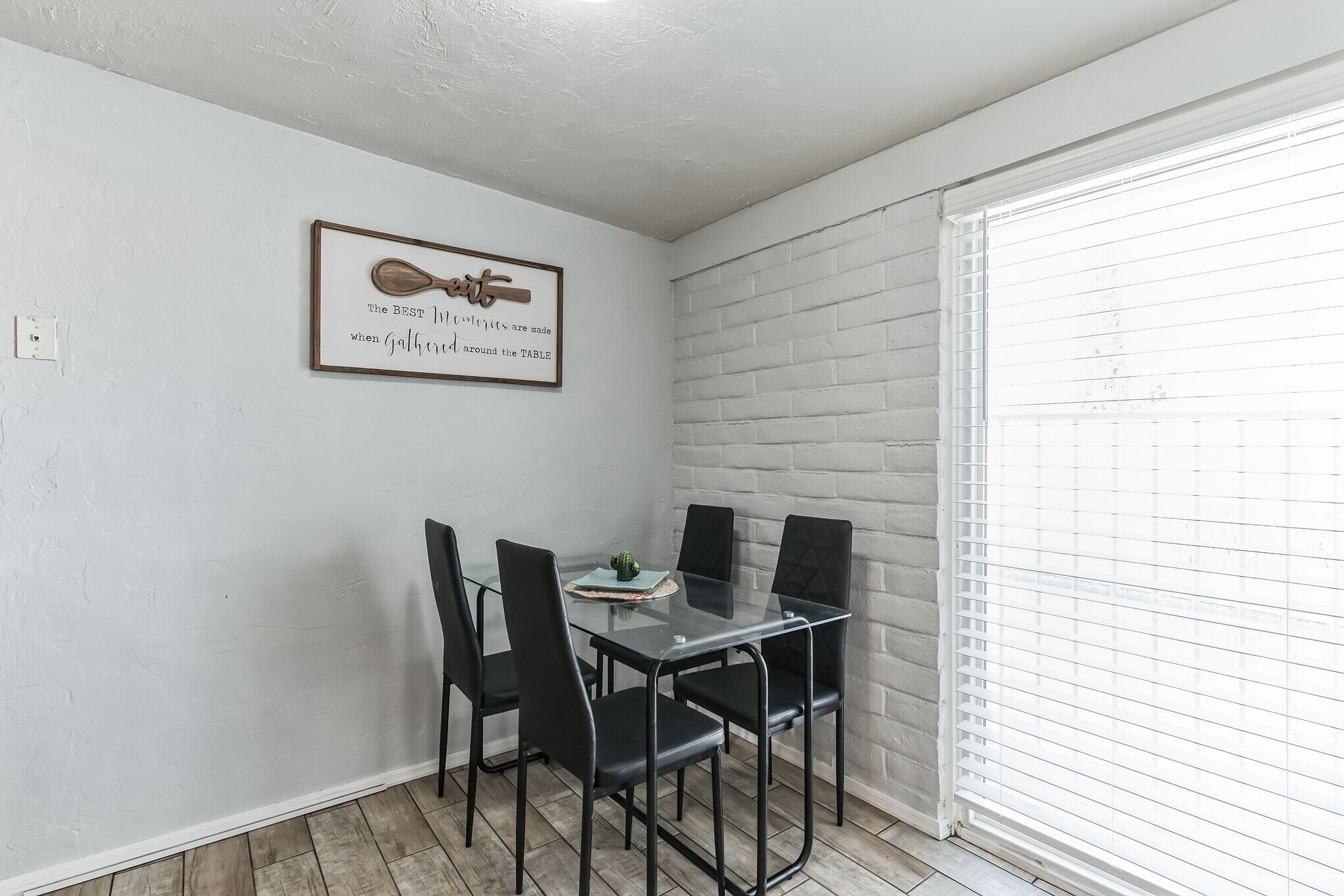 Un comedor con una mesa de vidrio y sillas negras, una cita enmarcada en la pared y un muro de ladrillo blanco.