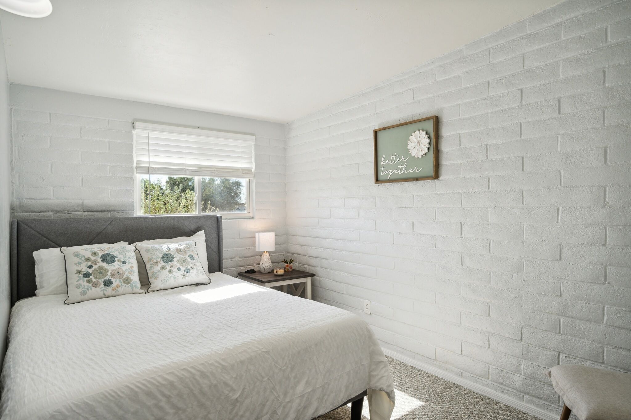 A bedroom with a white brick wall, a bed with floral pillows, a bedside table with a lamp, and a framed quote on the wall.