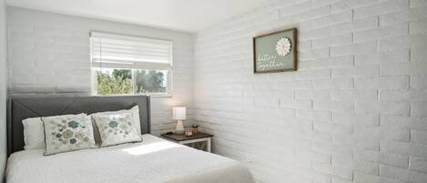 A bedroom with a white brick wall, a bed with floral pillows, a bedside table with a lamp, and a framed quote on the wall.