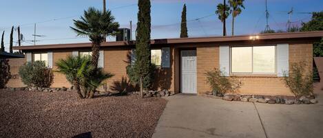 A single-story building with a brick facade, a front door, and a small garden area with rocks and plants.
