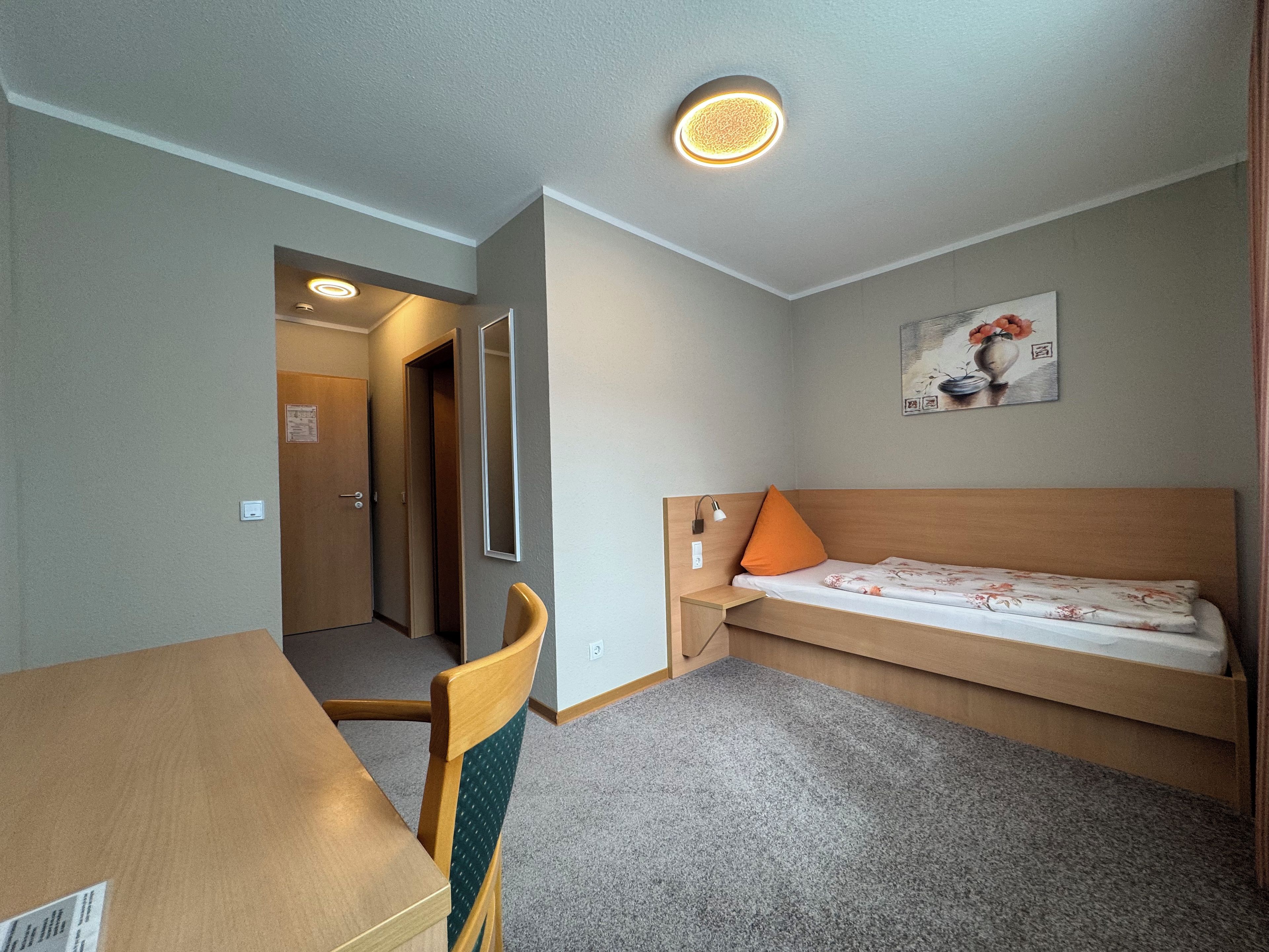 A small, neatly arranged room with a single bed, a desk, and a chair.
