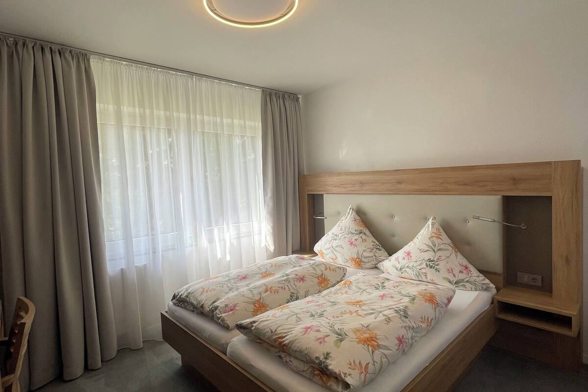 Ein Doppelbett mit Blumenmuster auf der Bettwäsche, ein Holz-Kopfstück, ein Nachttisch und eine Deckenleuchte.