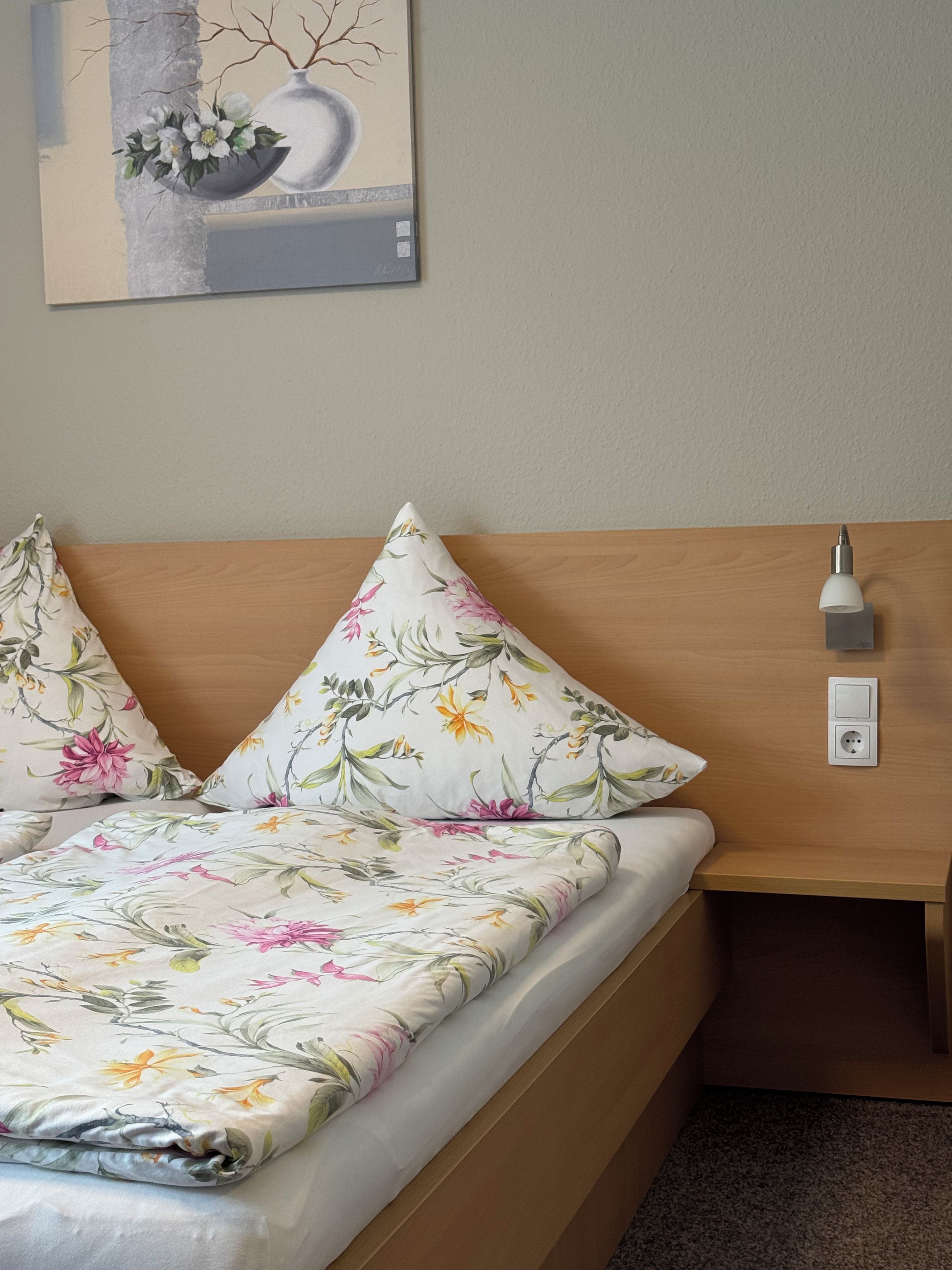 Ein Bett mit floraler Bettwäsche, ein Nachttisch, eine Wandlampe und ein Gemälde über dem Bett.