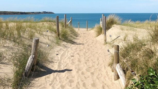 Ein sandiger Küstenpfad führt zum Meer, begleitet von Holzbalken und Gras an beiden Seiten.