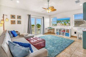 A modern living room with a sofa, ottoman, and a large rug. There is a ceiling fan, a bookshelf, and a TV mounted on the wall.