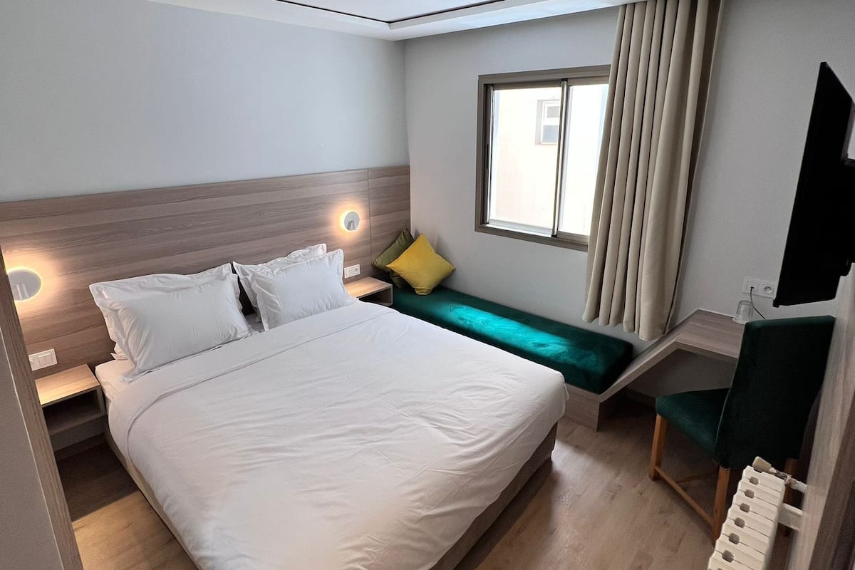A hotel room with a large bed, bedside tables, a desk, a chair, a window with curtains, and a flat-screen TV mounted on the wall.
