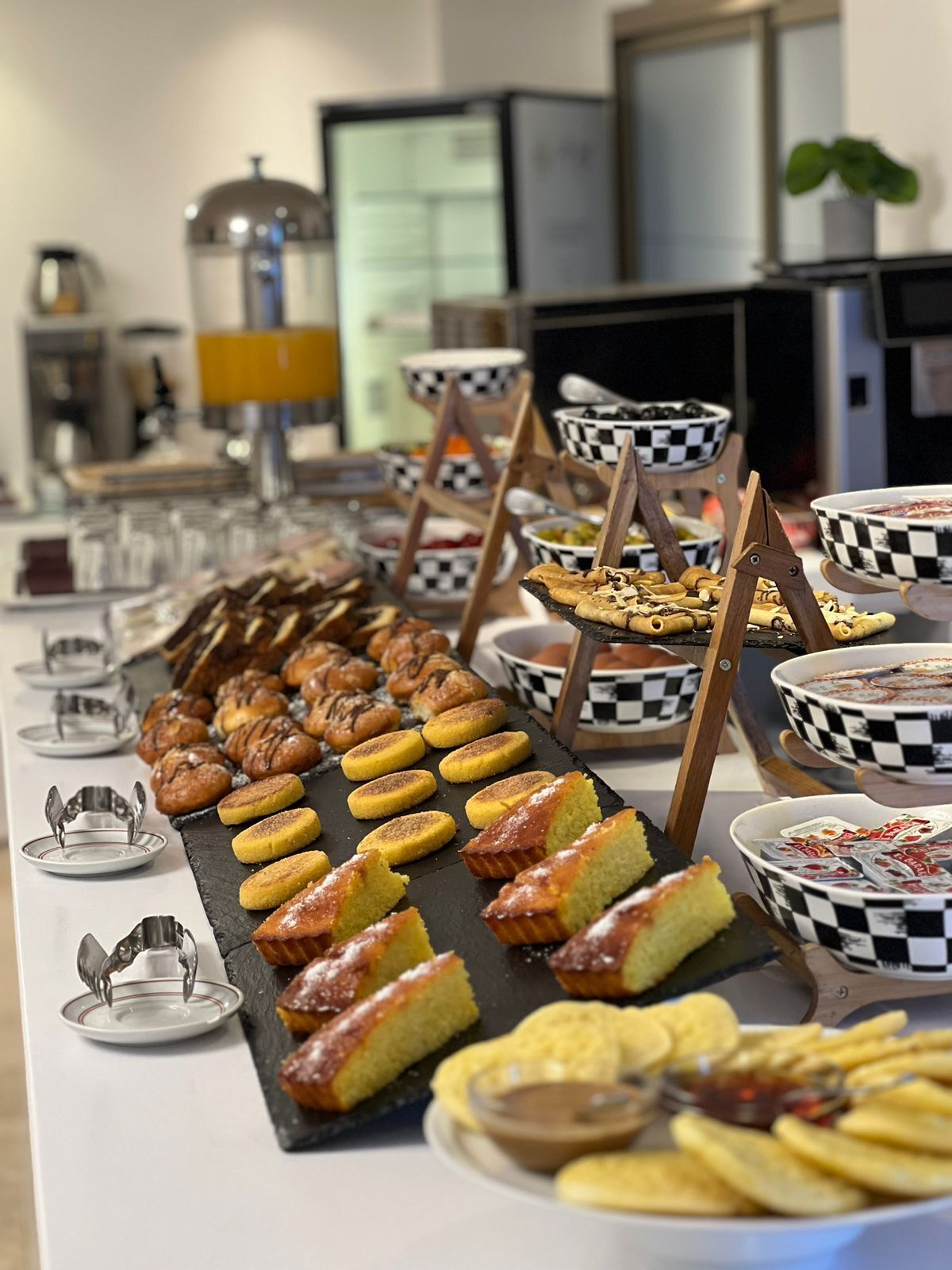A buffet table with various baked goods and pastries, including muffins, cookies, and bread, arranged on a long table with checkered bowls and serving utensils.