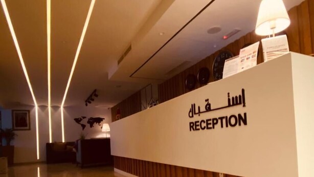 A reception area with a modern design, featuring a reception desk labeled "RECEPTION" and a sign with Arabic text. - Hotel Al Fajr (Oujda)
