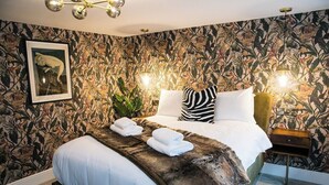 Une chambre à coucher avec un papier peint à motifs floraux, un lit recouvert de draps blancs, deux lampes de chevet et un tableau encadré au mur.