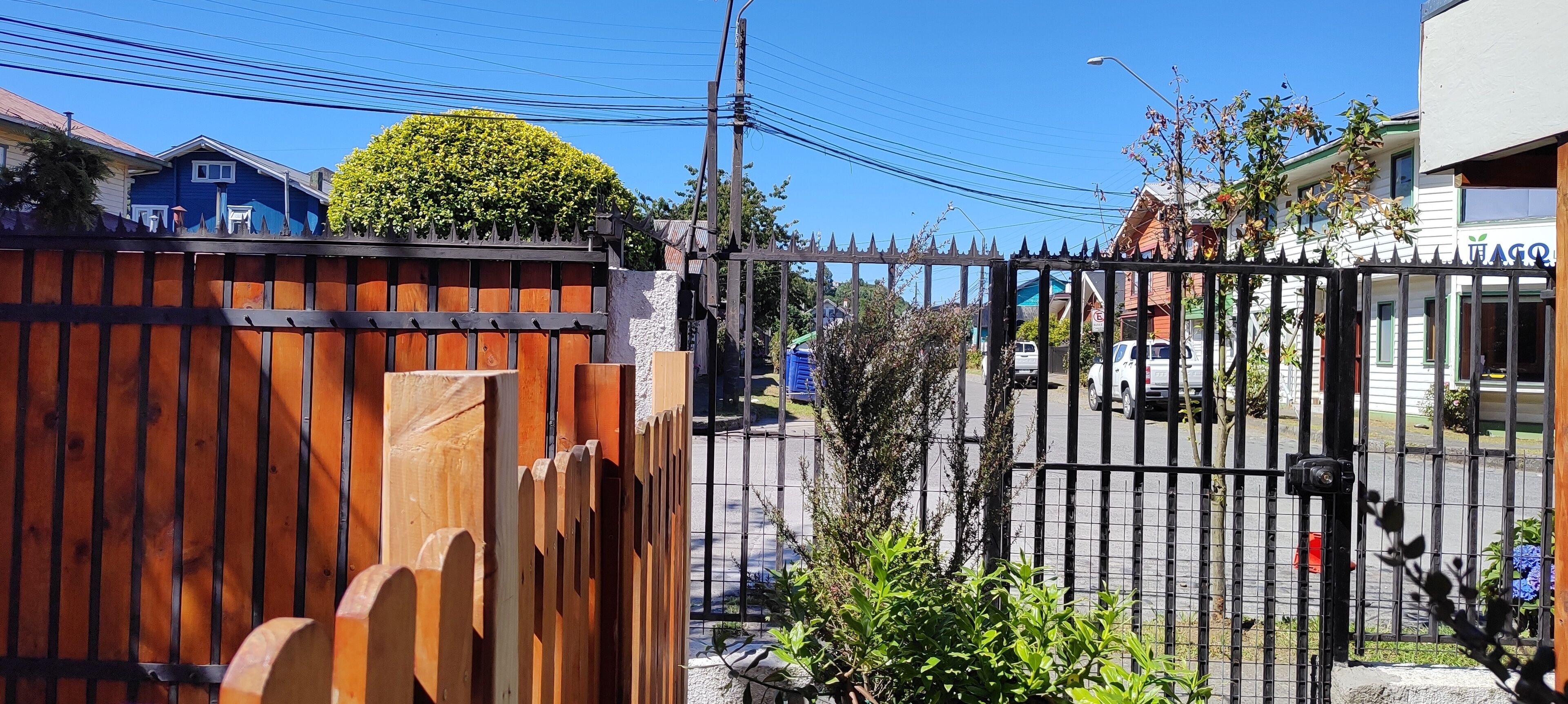 Un área cercada con valla de madera y portón metálico, un edificio azul y un edificio blanco con un letrero.