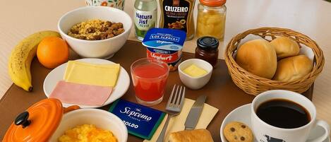Una mesa de desayuno con diversas opciones de comida, incluyendo un tazón de cereal, un vaso de jugo, un plato de jamón, queso y una cesta de panecillos.