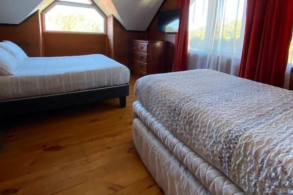 Ein Schlafzimmer mit zwei Betten, Holzboden, schrägen Wänden, einem Fernseher und einem Fenster mit Vorhängen.