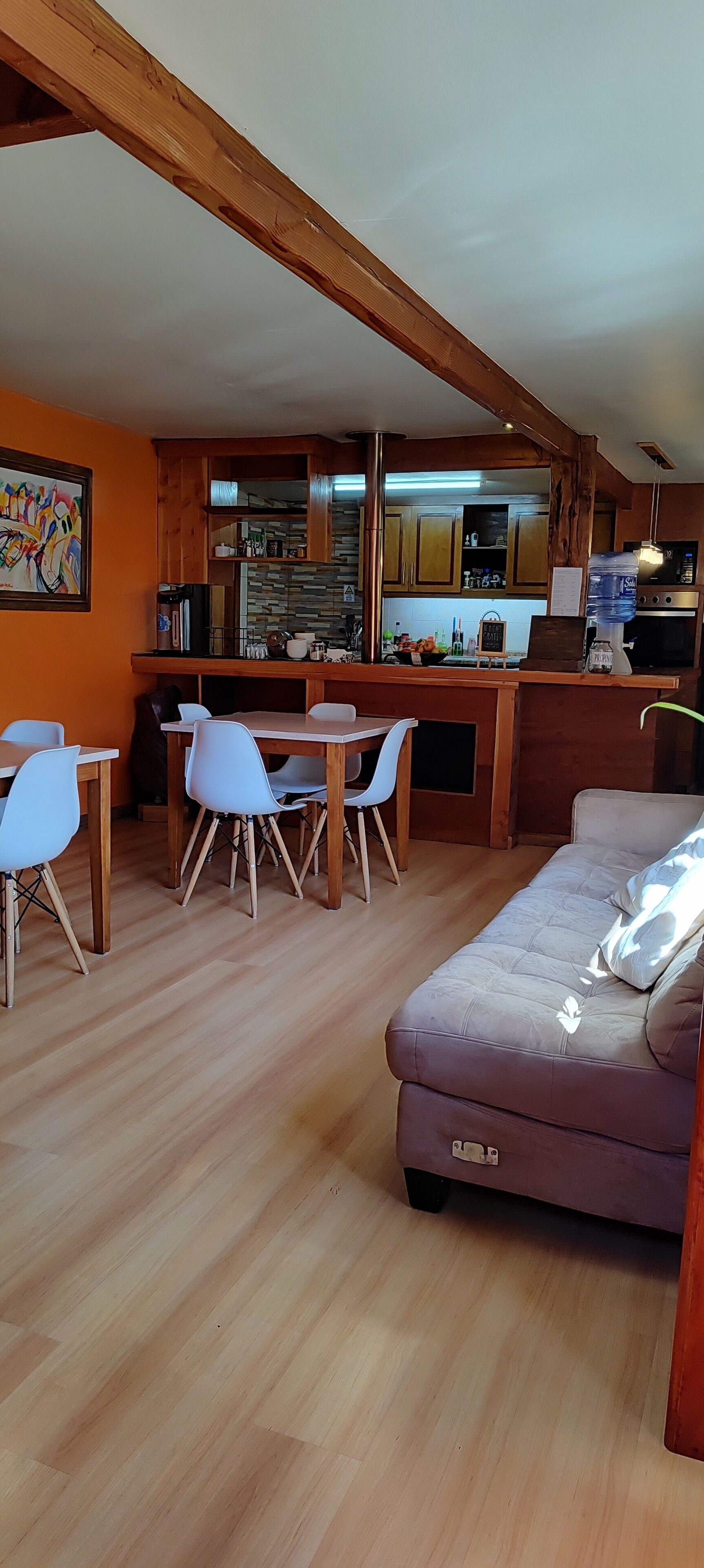 Amplia sala de estar con sofá, mesa de comedor y sillas. Se ve una cocina con armarios de madera y un dispensador de agua al fondo.