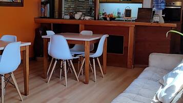 Amplia sala de estar con sofá, mesa de comedor y sillas. Se ve una cocina con armarios de madera y un dispensador de agua al fondo.