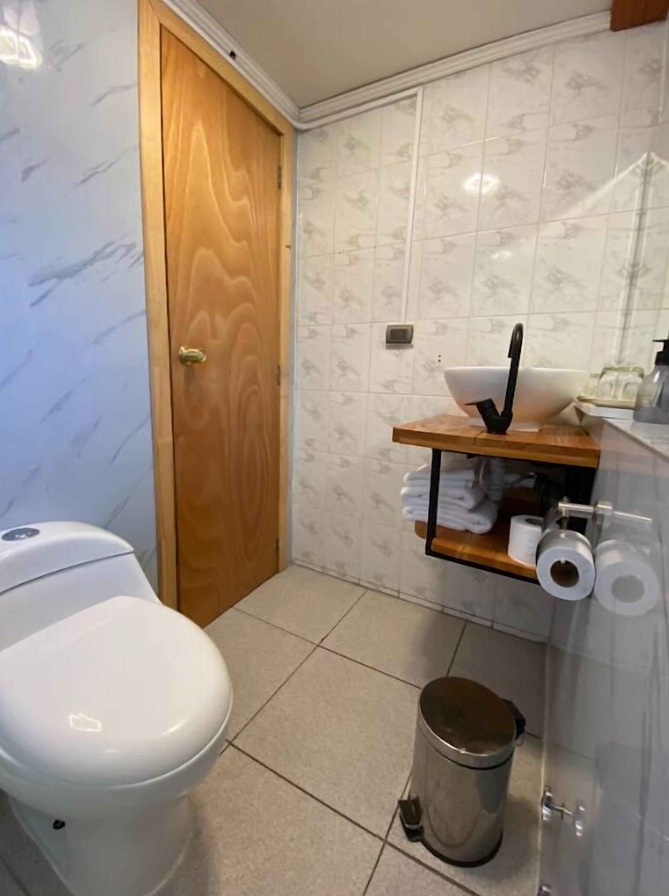 A small bathroom with a toilet, a wooden door, a sink with a mirror, and a trash can.