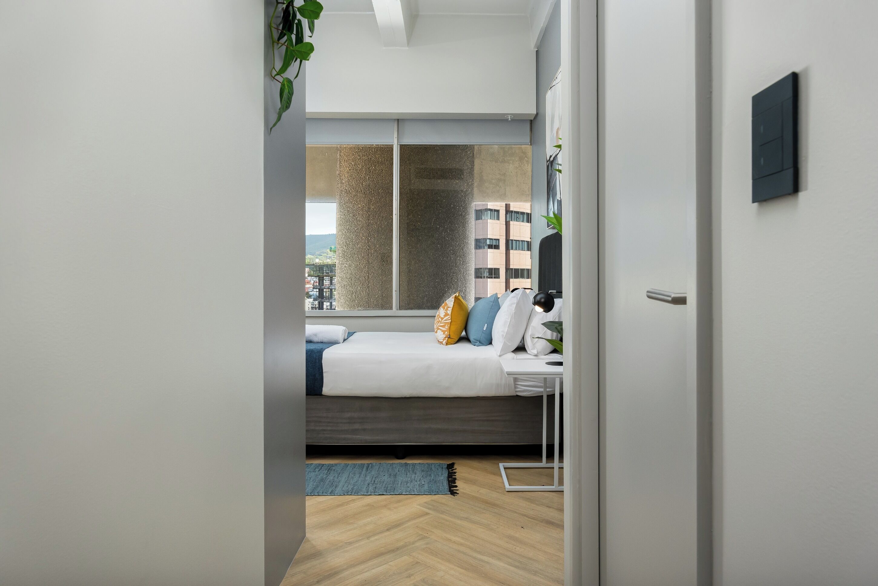 A modern bedroom with a bed, bedside table, and a view of the city through a large window.