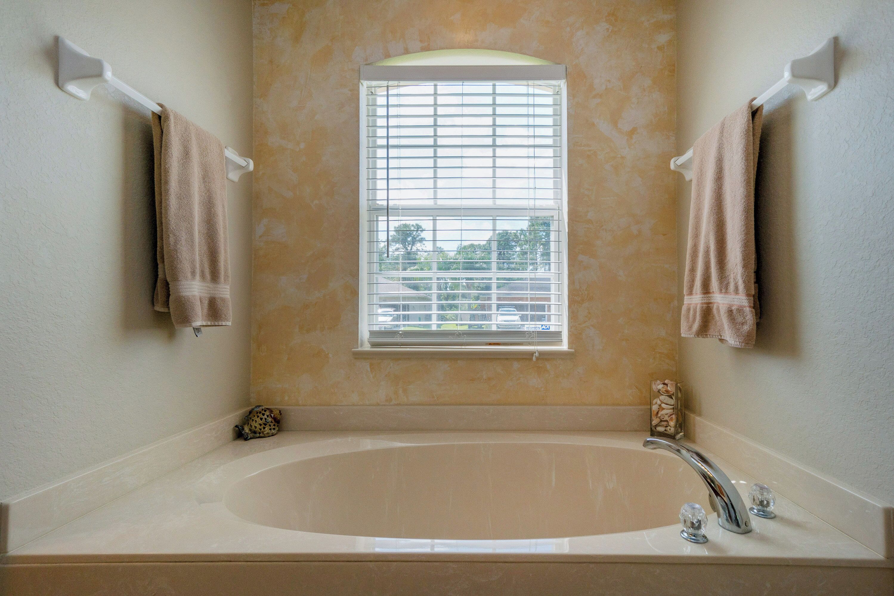 Une salle de bain avec une grande baignoire, une fenêtre avec des stores et deux serviettes suspendues à des porte-serviettes.