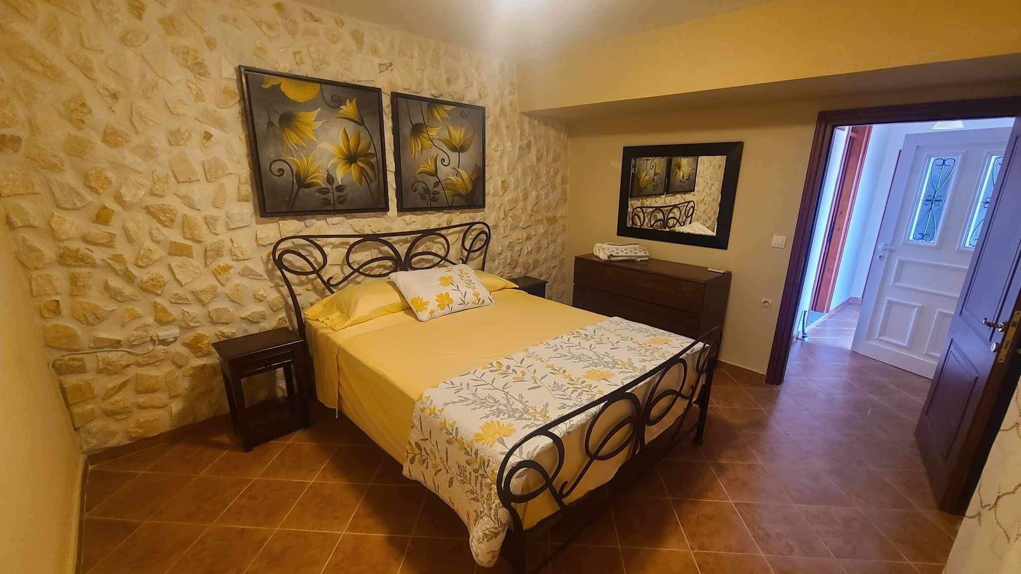 A bedroom with a stone wall, a bed with a floral bedspread, two nightstands, and a door leading to another room.