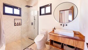 A modern bathroom with a glass-enclosed shower, a toilet, a sink with a wooden vanity, and a mirror with a round frame. - Villa Damiana Adults Only (El Pescadero)
