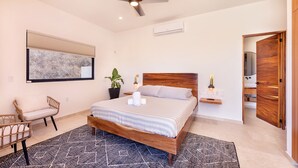 A modern bedroom with a wooden bed, a large window with blinds, a potted plant, a bedside table with a lamp, and a chair. - Villa Damiana Adults Only (El Pescadero)