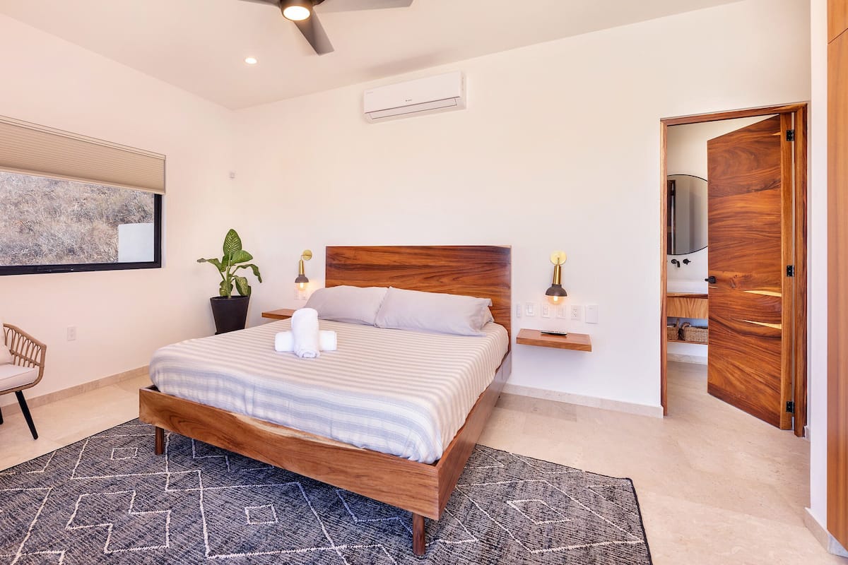 Un dormitorio moderno con una cama de madera, un ventanal con persianas, una planta en maceta, una mesita de noche con una lámpara y una silla.