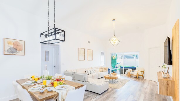 A spacious living room with a large sofa, a dining area with a wooden table, and a TV mounted on the wall. - Encanto /12 Ppl/ Pool/ Close to Disney Parks/ Games (Clermont)