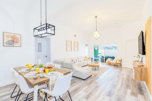 A spacious living room with a large sofa, a dining area with a wooden table, and a TV mounted on the wall. - 5BR Family Pool Home Near Disney Game Room  Sleeps 12 (Clermont)