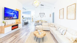 A modern living room with a flat-screen TV, a wooden entertainment unit, a beige sofa, and a round coffee table. - Encanto /12 Ppl/ Pool/ Close to Disney Parks/ Games (Clermont)