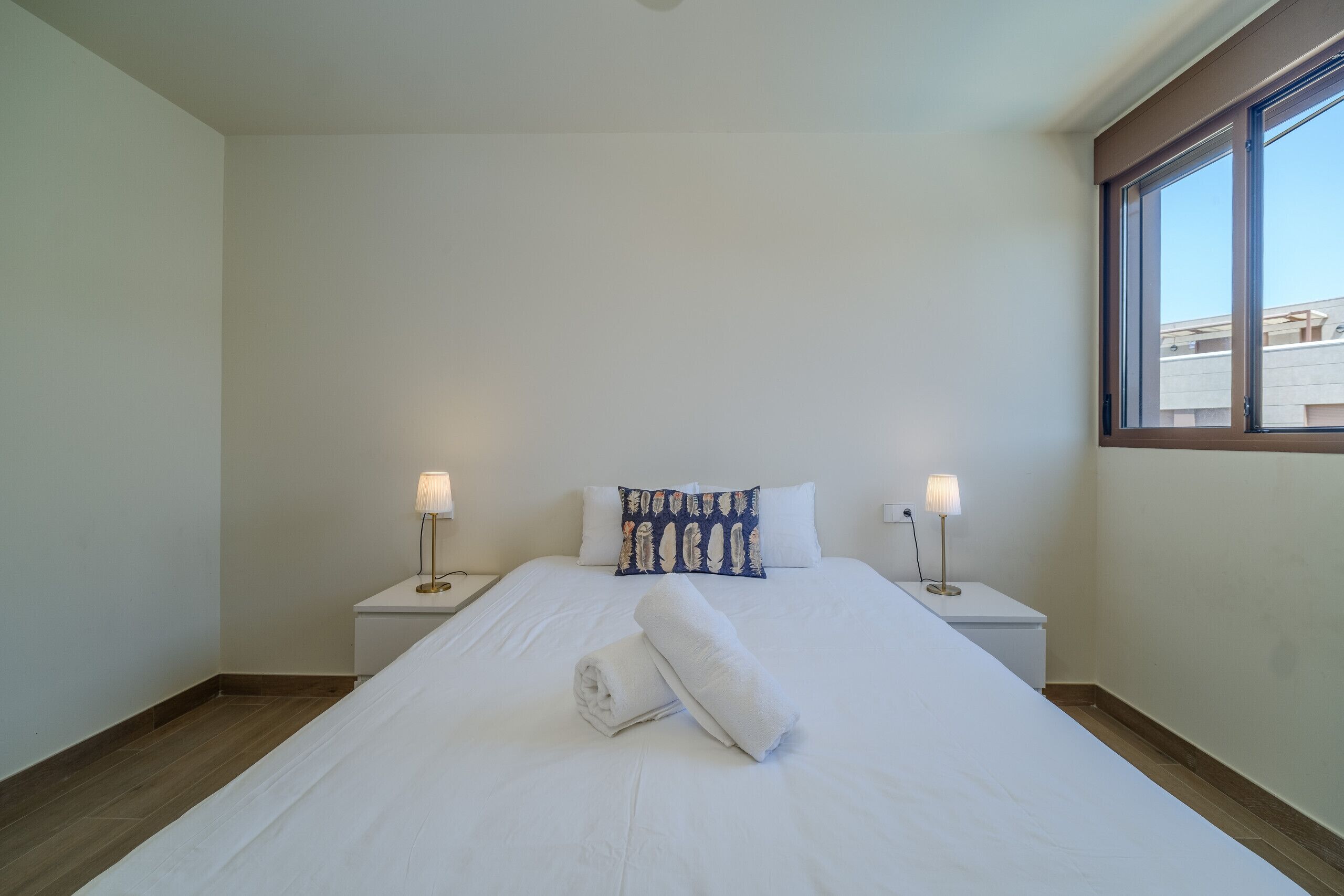 A neatly made bed with a rolled towel, two bedside lamps, and a pillow with a leaf pattern.