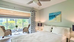 Un dormitorio con una cama grande, dos sillas de mimbre, una mesita, un ventilador de techo y un cuadro de una escena de playa.