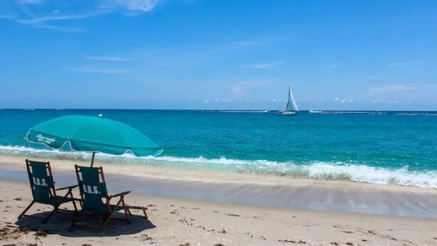Una playa con dos sillas de playa y un paraguas turquesa, un velero a lo lejos y un cielo azul despejado.