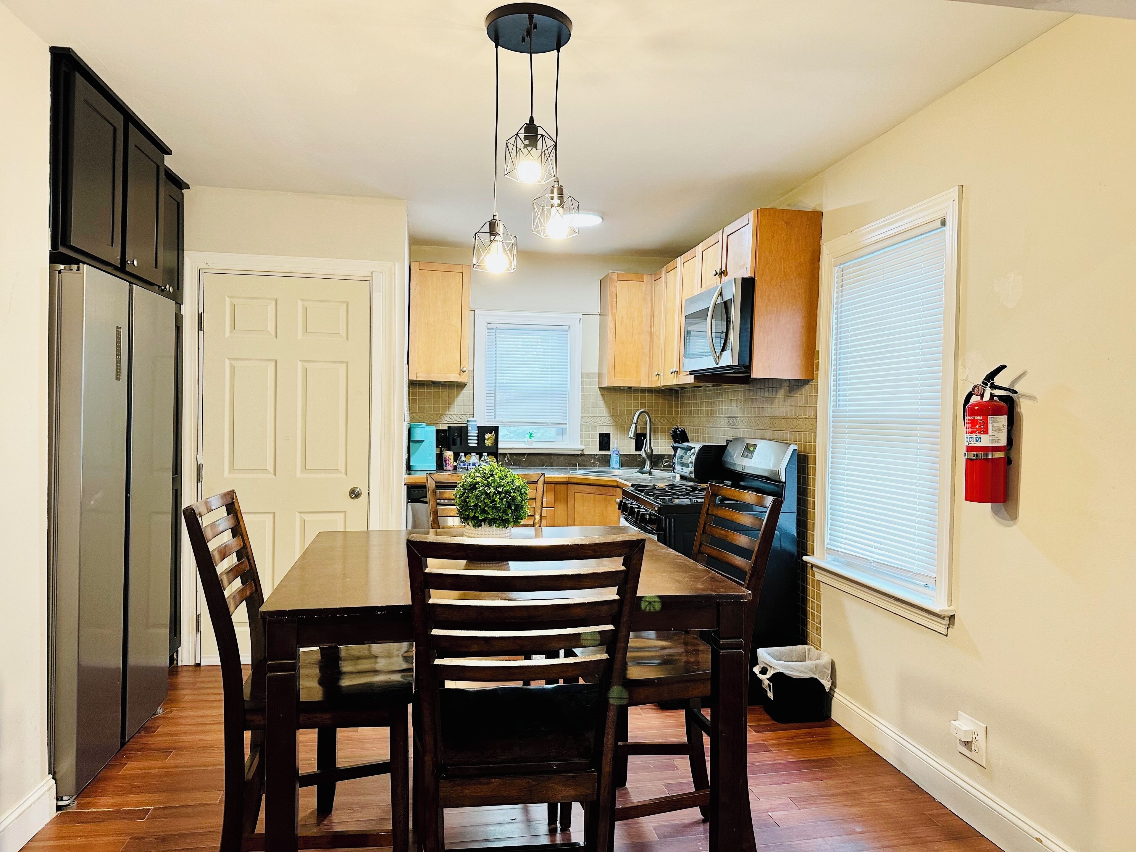 A kitchen with wooden cabinets, a stainless steel refrigerator, a microwave, and a stove. There is a dining table with chairs and a hanging light fixture.