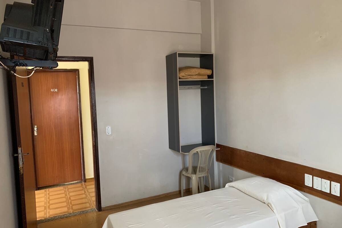 A small, simple room with a bed, a desk with a chair, a shelf with a folded blanket, and a television on a stand.