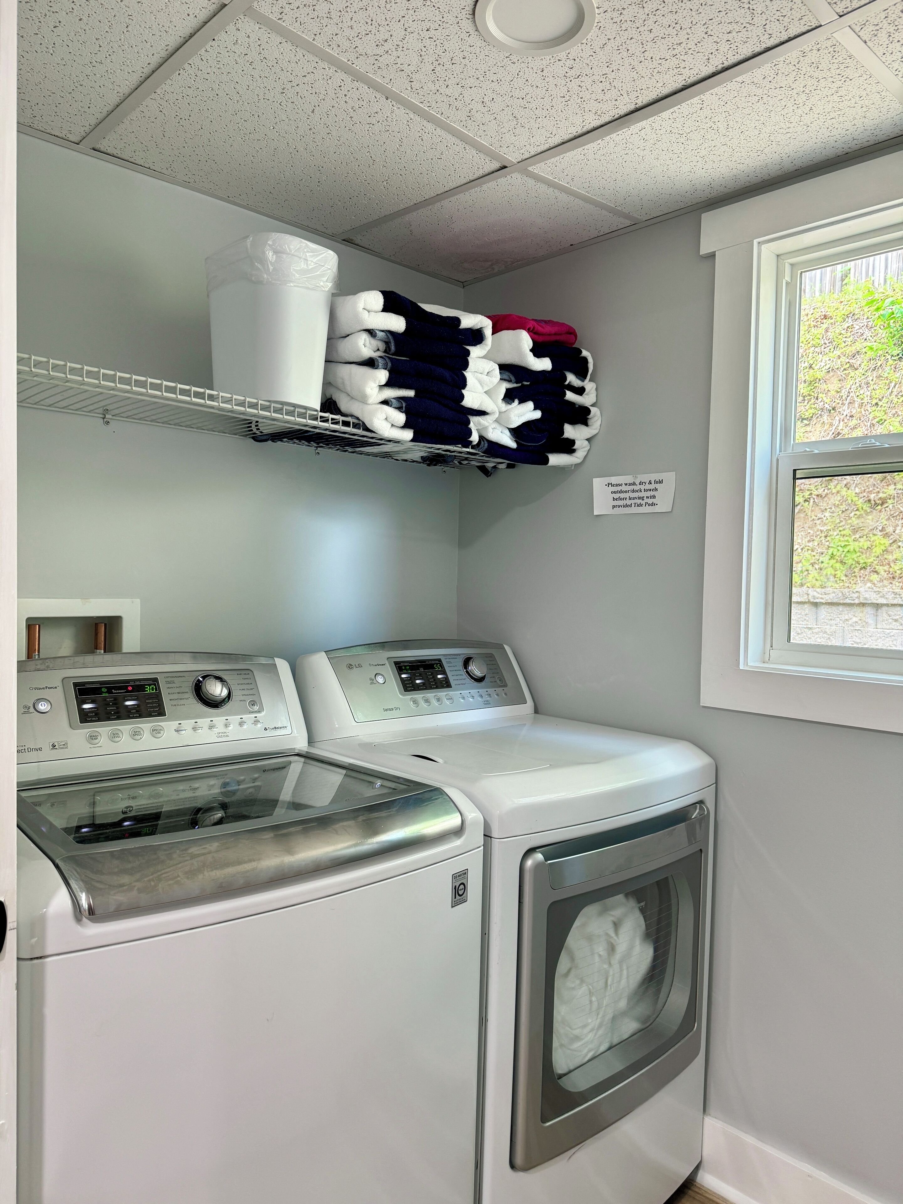 A laundry area with two washing machines, a drying rack, and a window with a view of greenery.