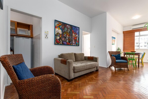Una sala de estar con un sofá, dos sillas de mimbre, una mesa de comedor con sillas, un microondas y un refrigerador.