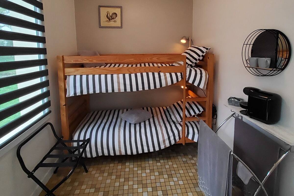A small, compact room with a bunk bed, a desk with a chair, a small table, and a wall-mounted mirror.